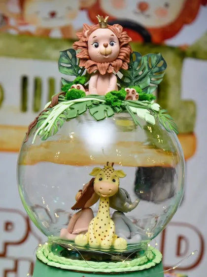 A stunningly detailed jungle-themed cake topper, featuring a lion king sitting atop a glass globe with other safari animals inside.