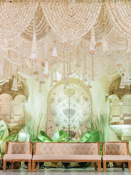 An opulent stage design featuring a canopy of cascading white flowers and intricate details. The backdrop includes traditional golden jharokhas and lush greenery, creating a setting that is both regal and deeply romantic.
