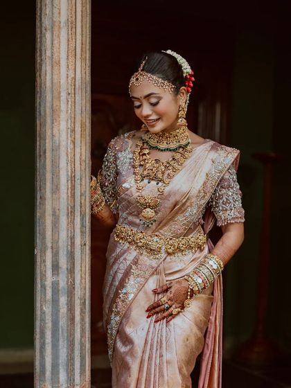A bride in a beautiful pastel saree, her intricate jewelry and serene smile captured against a stone pillar.