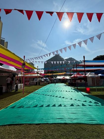 A long shot of the main walkway at Kukdukoo Pune, showing the scale of the event. The green turf path guides visitors through rows of vendor stalls, all sheltered under the colorful bunting and canopies I installed.