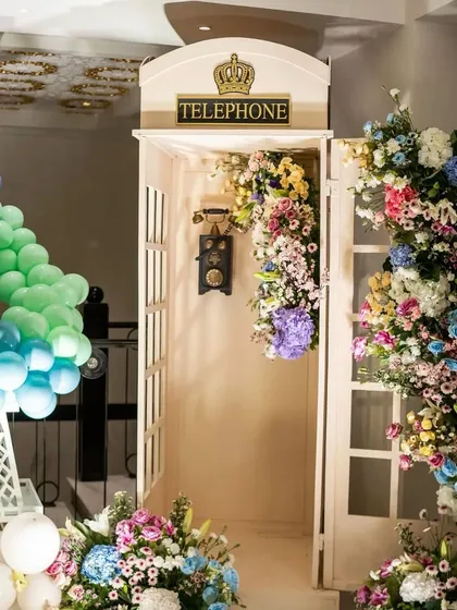 A whimsical and elegant photo booth created for a Parisian baby shower, featuring a classic white telephone booth overflowing with a stunning arrangement of colorful flowers. This created a perfect, memorable spot for guest photos.