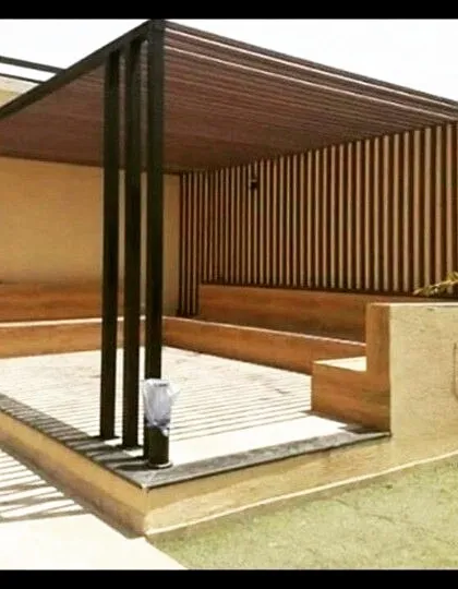 This pergola features a dark metal frame with a slatted roof and back wall, paired with built-in wooden bench seating. The design creates a defined, shaded seating area within a larger garden.