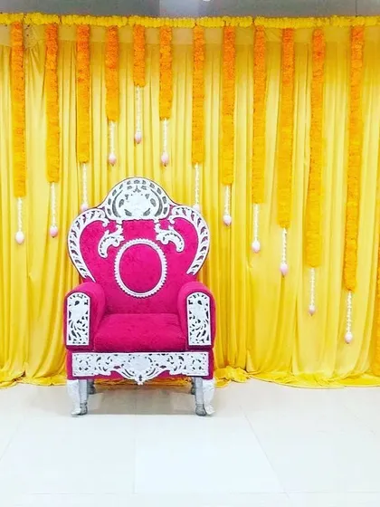A simple and elegant backdrop using yellow drapes and hanging marigold garlands. The ornate pink chair adds a pop of color and a royal touch.