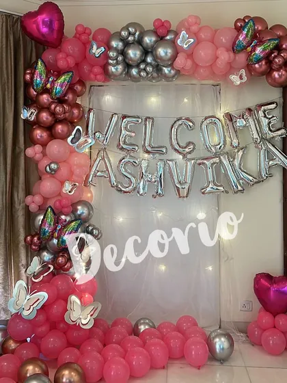 A welcoming home decoration for baby Ashvika. The setup features a 'WELCOME' foil balloon banner and a beautiful arch of pink, rose gold, and silver balloons with butterfly accents.