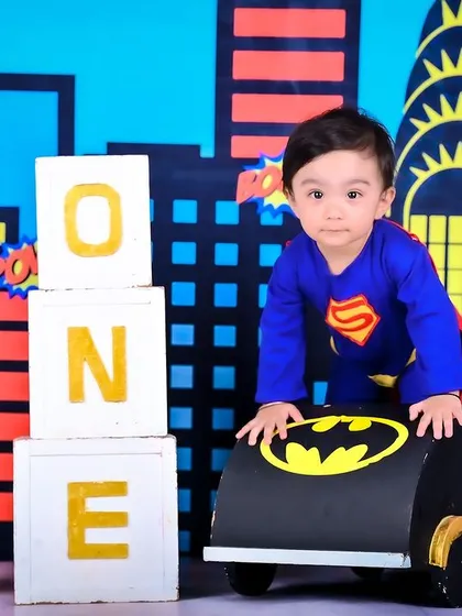A superhero-themed first birthday shoot, with the baby dressed as Superman next to a Batmobile prop.