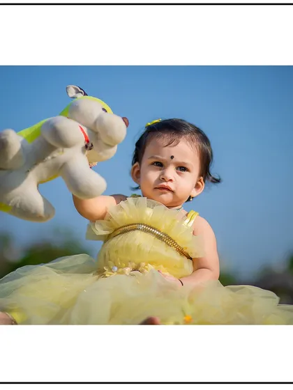 A beautiful close-up of a baby girl in a lovely yellow dress, playing with her stuffed toy during an outdoor session under a clear blue sky.