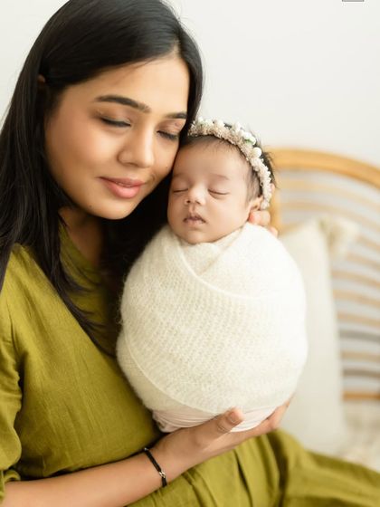 A serene portrait of a mother holding her peacefully sleeping, swaddled newborn. The olive green of her dress adds a touch of color to the neutral setting.