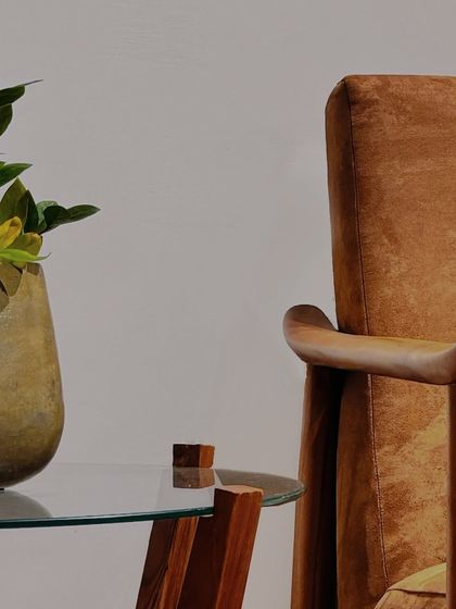 A detail of a suede-upholstered armchair next to a glass-topped side table. The smooth, sculpted teak armrest is a key feature, designed for both beauty and comfort.