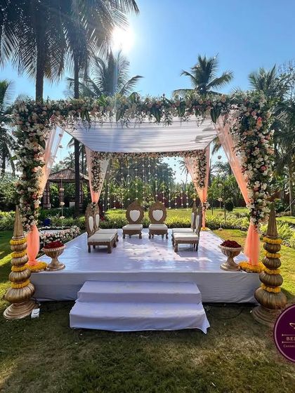 An elegant outdoor 'Muhurtham Mantap' set against a beautiful backdrop of palm trees. The simple white stage, pastel drapes, and fresh floral arrangements create a serene and picturesque setting for the wedding ceremony.