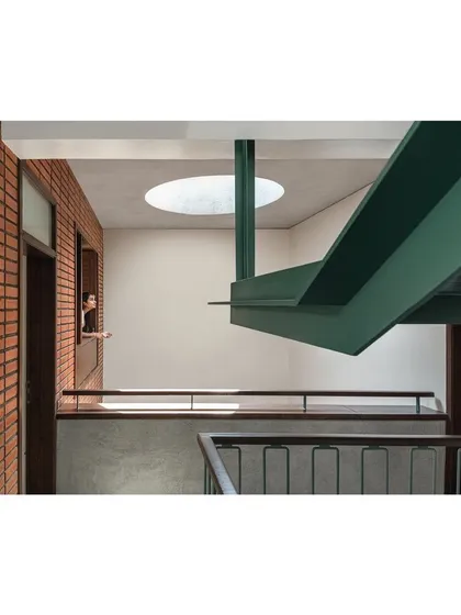 The main staircase is a central sculptural element within the Nairy Residence. A bold, green-painted steel beam supports the structure, creating a visual contrast with the raw brick wall and drawing the eye upwards towards a circular skylight, which bathes the double-height volume in daylight.