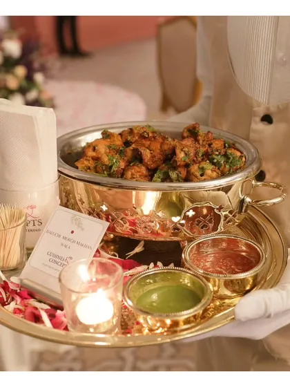 A tray of Custom Murgh Malai Tikka served in a beautiful brass dish with green and red chutneys. The presentation is elevated by the luxurious serveware and scattered rose petals.