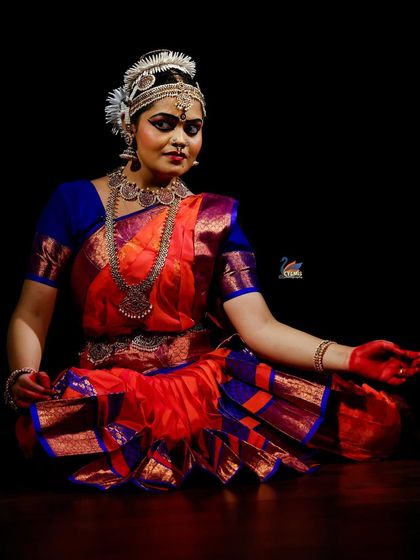 A seated portrait of a Bharatanatyam dancer, showcasing her intricate costume and makeup against a simple black backdrop for a striking effect.