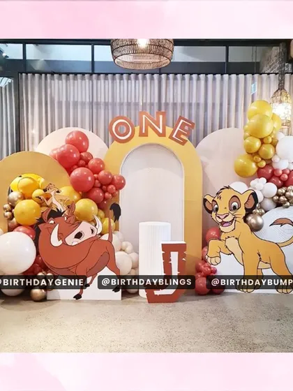 A playful Lion King first birthday setup with large character cutouts, layered arch backdrops, and a fun balloon color scheme of red, yellow, and white.