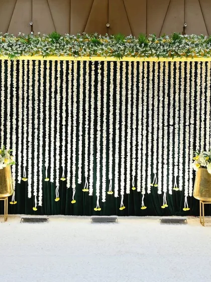 A simple yet elegant Mehendi backdrop with strings of white flowers against a dark green curtain, framed in bright yellow.