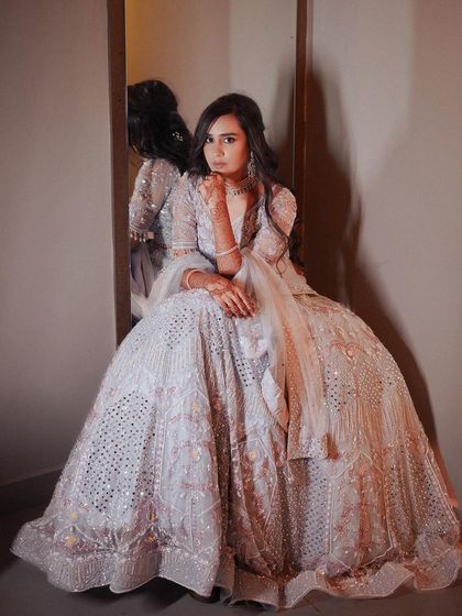 A full-length portrait of the bride in a stunning, voluminous white lehenga, seated in a corner.