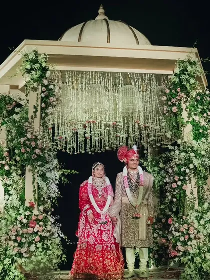 The couple stands together at the Varmaala mandap, framed by lush floral pillars and a sky filled with dramatic smoke effects. This moment captures the beginning of their journey in a setting designed for romance and splendor.