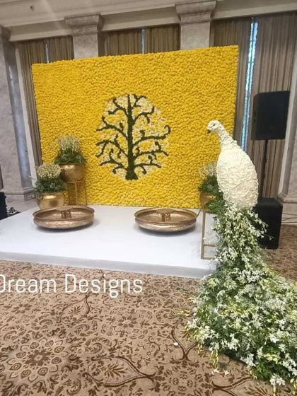 A gorgeous stage for a mehendi ceremony. The backdrop is a solid wall of yellow flowers featuring a tree-of-life motif, complemented by a stunning peacock sculpture made of white flowers.