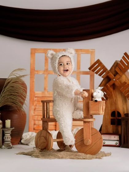 A happy toddler in a fluffy bear costume stands on a wooden tricycle in a rustic studio setup with a miniature windmill.