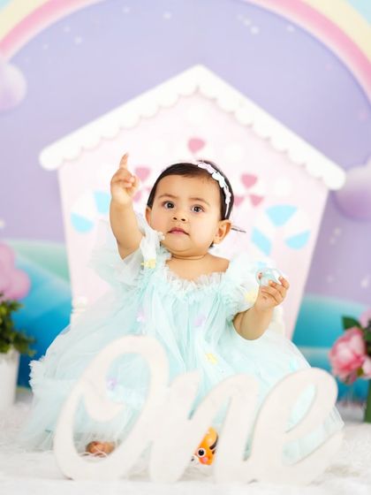 A sweet baby girl in a pastel blue dress points up during her first birthday shoot, set against a whimsical rainbow backdrop.