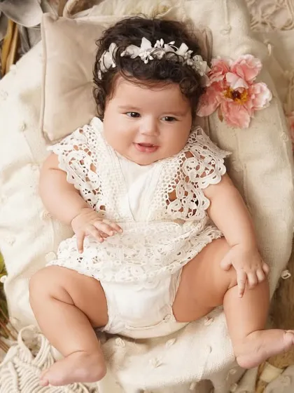A full-body shot of a happy baby girl in a lovely lace outfit, posed comfortably in a boho-styled basket with floral accents.