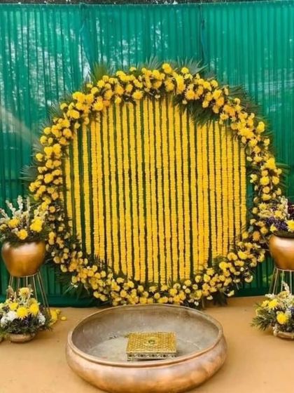 A classic Haldi backdrop for outdoor functions. This setup features a large circular frame filled with strings of yellow marigolds, a green panel background, and a traditional brass urli in the center for the ceremony.