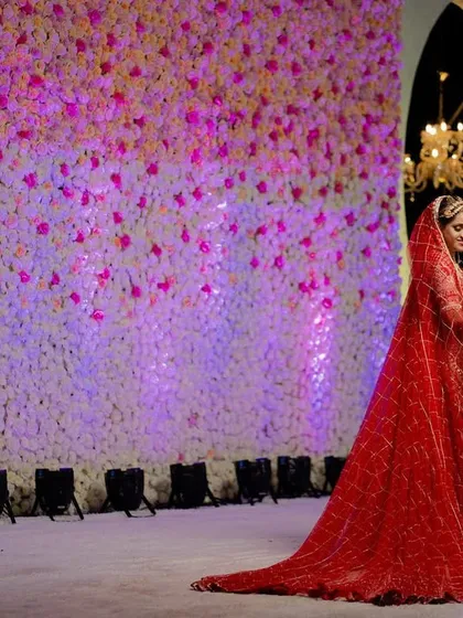 A grand and dramatic wedding portrait. The bride's long, flowing veil against the wall of flowers creates a stunning visual.