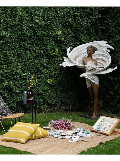 A full view of the garden setting with the elegant sculpture of a woman. The scene is set for a creative afternoon, with art supplies laid out on a mat, suggesting the landscape is a source of inspiration.