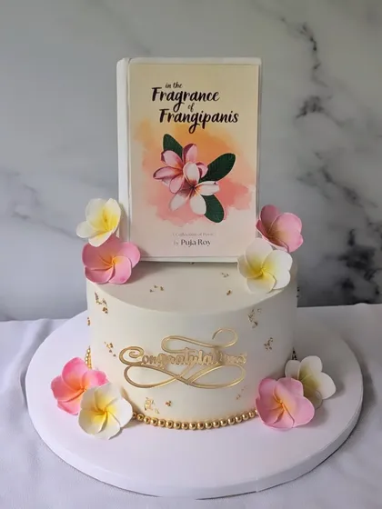 A beautiful and elegant cake designed for a book launch. The centerpiece is an edible replica of the book cover for "In the Fragrance of Frangipanis," surrounded by handcrafted sugar flowers that match the theme.