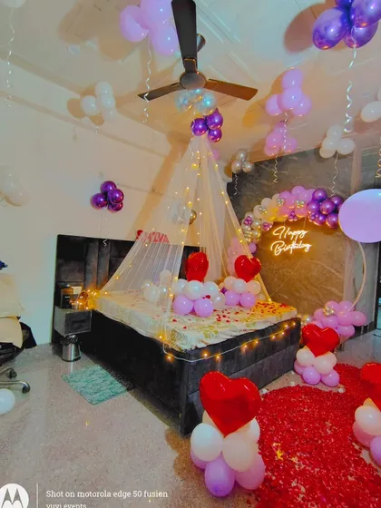A dreamy birthday setup in purple and white, featuring a light-up canopy over the bed. The room includes a balloon arch with a 'Happy Birthday' neon sign and a path of red rose petals on the floor.