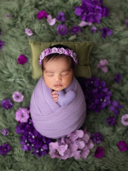 An overhead shot of a swaddled baby holding a tiny purple heart, surrounded by beautiful flowers.