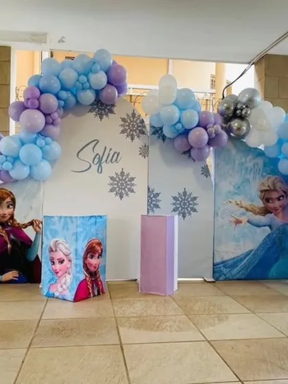 A wide view of a Frozen-themed 5th birthday party, showcasing a large balloon arch over multiple backdrops with characters and a light-up marquee number.