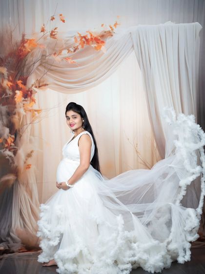 A mother-to-be looking ethereal in a white gown with a dramatic flowing train, set against an autumn-themed boho backdrop.