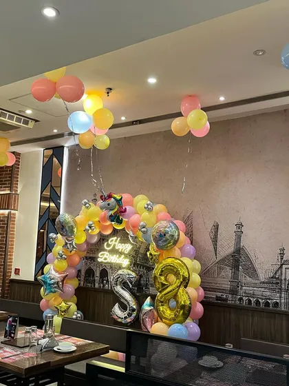A unicorn-themed balloon arch set up over a table in a restaurant, featuring a neon sign and foil balloons for the age '8'.