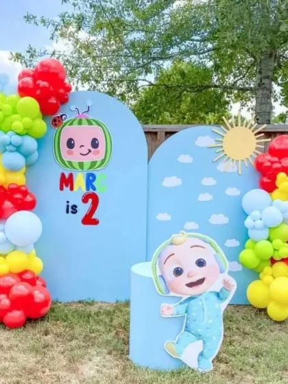 An adorable Cocomelon-themed outdoor party for a 2nd birthday. The setup uses two blue arched backdrops to create a sky scene, with colorful balloon clusters and a JJ cutout.
