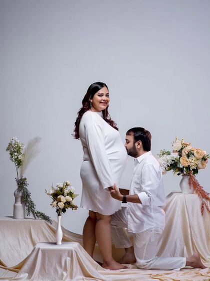 A classic pose filled with anticipation and love. The father-to-be kisses the baby bump, a gesture of affection for both mother and child. The all-white theme and floral arrangement create a pure and elegant setting.