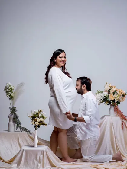 A classic pose filled with anticipation and love. The father-to-be kisses the baby bump, a gesture of affection for both mother and child. The all-white theme and floral arrangement create a pure and elegant setting.