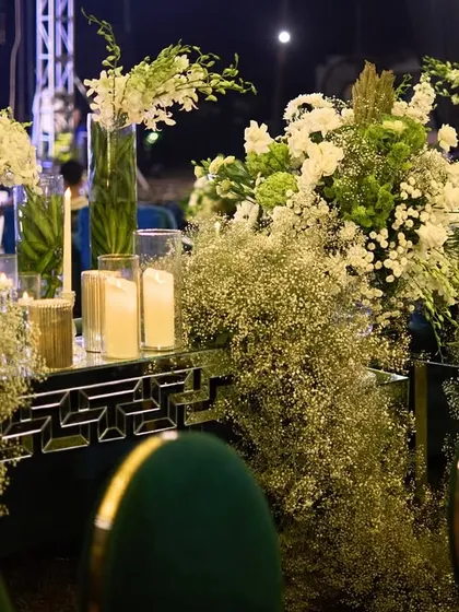 A tablescape from a Sangeet night, featuring a mirrored table runner overflowing with baby's breath and white flowers, set against plush teal chairs.