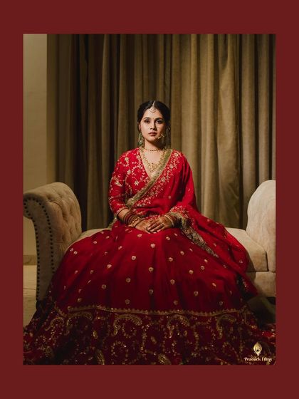 A regal bridal portrait. The bride, seated in her magnificent red lehenga, looks directly at the camera with grace and confidence.