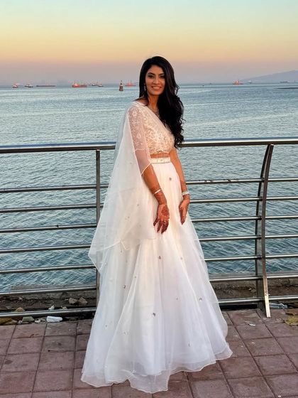 This full-length shot captures the complete look against a beautiful seaside backdrop. The client's white lehenga and natural, glowing makeup are perfect for a destination wedding celebration.