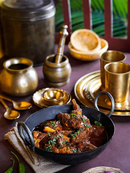 A bowl of authentic Maharashtrian mutton curry, styled with traditional brass kitchenware. The dark and moody lighting emphasizes the deep, rich colors of the gravy.