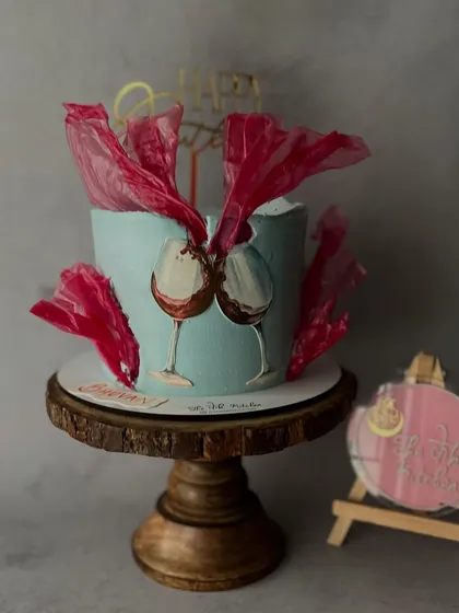A unique celebration cake with hand-painted wine glasses clinking in a toast. The design is set against a cool blue background and accented with abstract, vibrant red sugar sails for a modern, artistic flair.