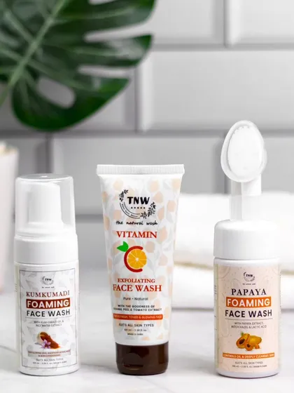 This bathroom counter scene features three face washes, including foaming and exfoliating types. The clean white tiles and a touch of green from a plant create a fresh, natural, and calming atmosphere.