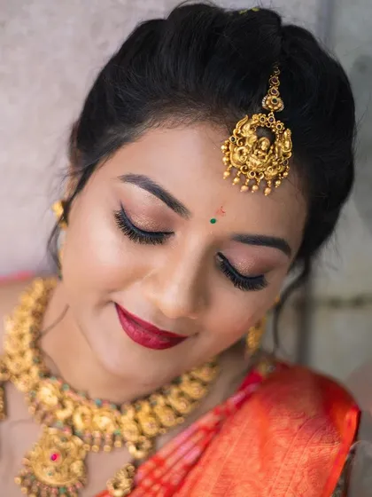 A stunning bridal portrait in a vibrant orange silk saree. This look features classic winged eyeliner and a bold red lip, paired with heavy temple jewellery for a truly traditional South Indian wedding style.