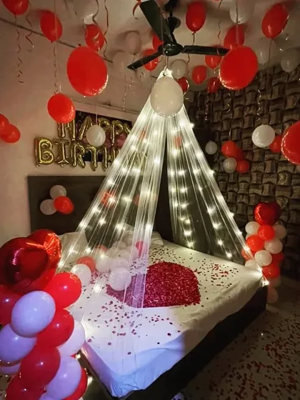A classic romantic bedroom decoration, perfect for birthdays or anniversaries. This popular design features a sheer canopy with fairy lights over the bed, a heart made of rose petals, and dozens of red and white balloons floating on the ceiling.