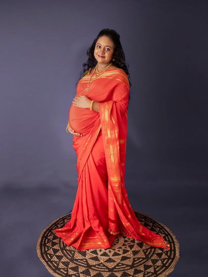 An elegant portrait in a vibrant orange saree. Standing on a rustic jute mat against a plain background, the focus remains entirely on the mom-to-be and her beautiful bump.