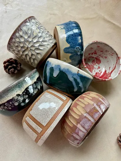 A collection of glazed student work, showing a range of styles and techniques. The pieces include a textured white cup, a speckled bowl, and a mug with a carved house design.