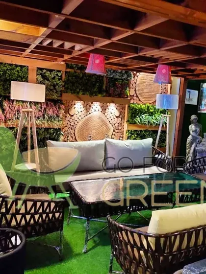 This image captures a complete outdoor lounge scene in our showroom. It features a dark-weave sofa set with light-colored cushions, set against a backdrop of vertical greenery and wood accents. The warm lighting from the floor lamps shows how you can create a cozy and inviting ambiance for evening relaxation.