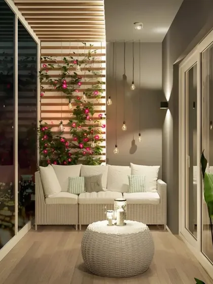 This elegant night-time view shows a comfortable white wicker sofa set against a slatted wood feature wall with climbing plants. The soft glow from pendant lights and the reflection from the glass doors create a peaceful and sophisticated atmosphere.
