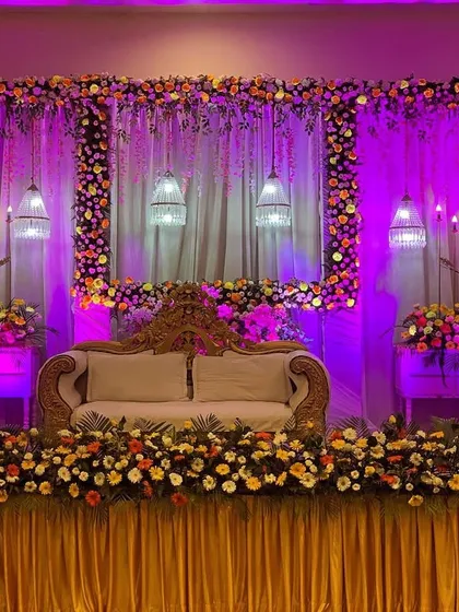 An elegant wedding stage design with a beautiful floral frame and dramatic purple uplighting. The combination of hanging birdcage lamps, candle stands, and a plush sofa creates a sophisticated and grand backdrop for the couple.
