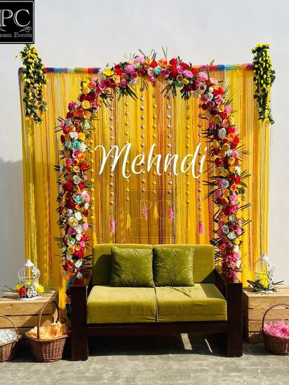 A bright and cheerful Mehndi backdrop. I used a vibrant yellow curtain as the base, adorned with a multi-colored floral arch and a simple 'Mehndi' sign for an elegant look.